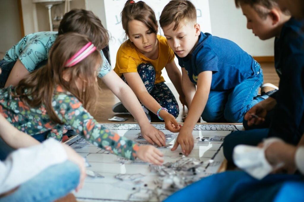 Grupa dzieci siedzi na podłodze wokół gry planszowej. Wyciągają ręce po karty leżące na planszy.