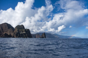 Widok na wody oceanu wokół Madery oraz na skały u jej brzegu. Nad oceanem kłębią się białe chmury. W oddali tęcza.