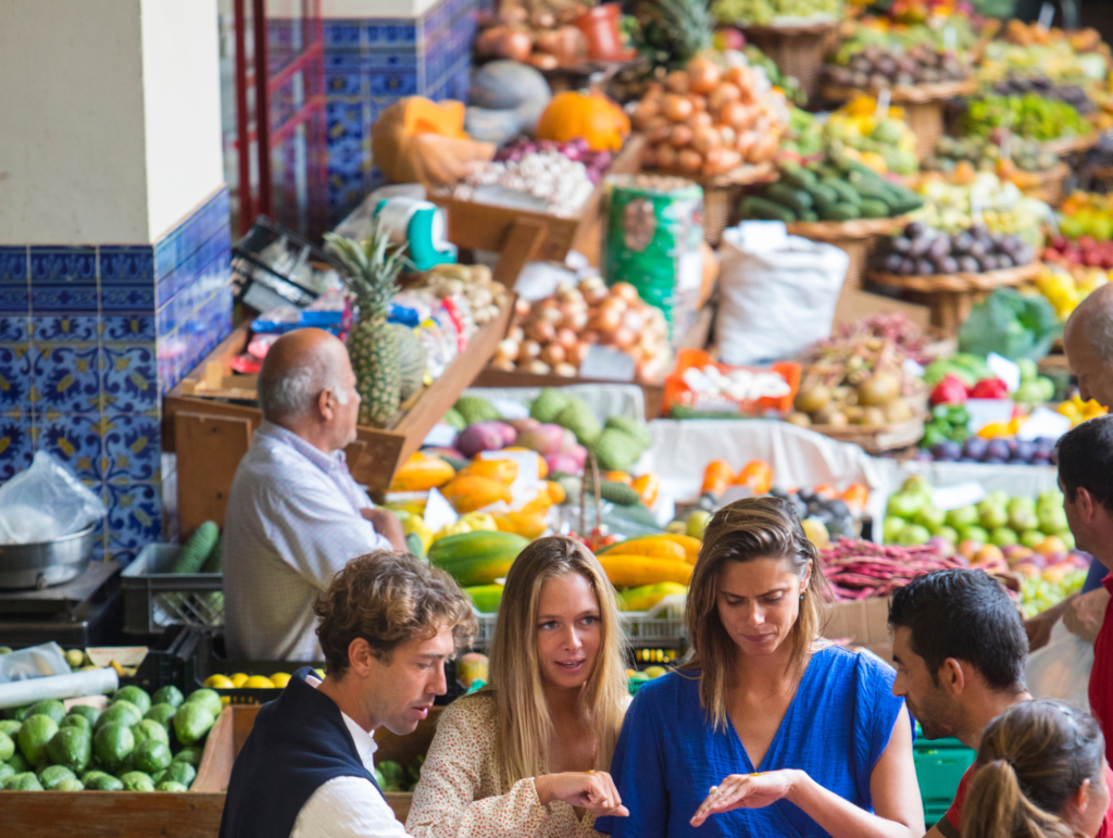 Targowisko na Maderze. Na straganach pełno kolorowych owoców i warzyw. Grupa ludzi robi zakupy.