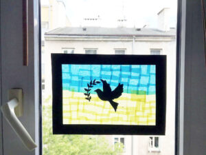 Gotowa praca wisie na oknie. To witraż z bibuły. Przedstawia gołąbka z gałązką oliwną na niebiesko-żółtym tle - fladze Ukrainy.