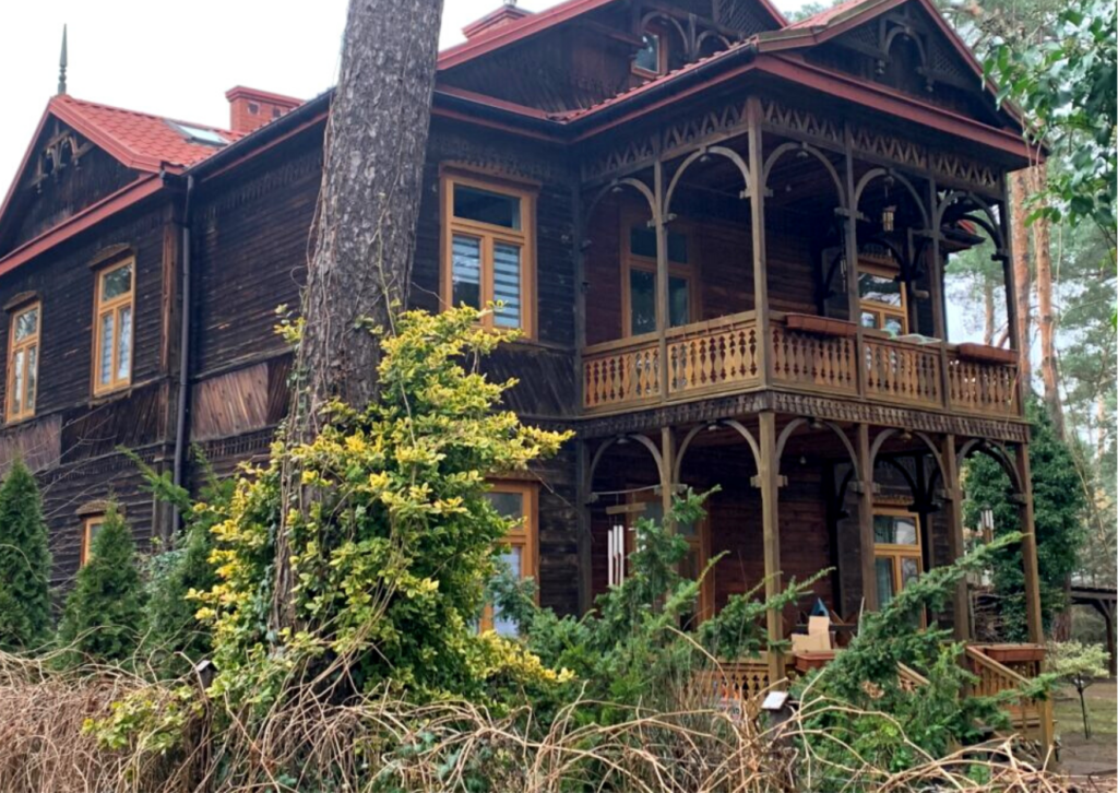 Pomiędzy drzewami stara drewniana willa z tarasem i balkonem z dekoracyjnymi tralkami.