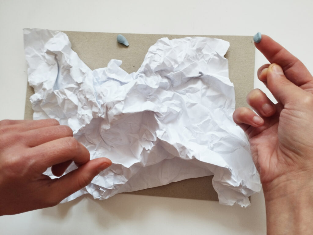 rzeźba terenu_zdjęcie 6 Ręce przyklejające zgniecioną kartkę papieru do szarego kartonu.