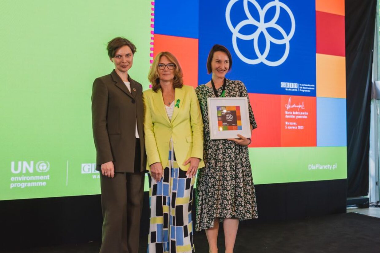 W centrum zdjęcia stoi trójka kobiet, z czego jedna po prawej stronie trzyma nagrodę.